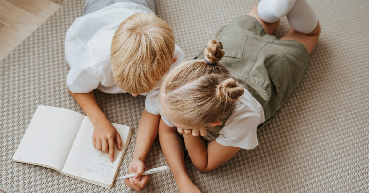 Två barn ligger på golvet och skriver i en bok