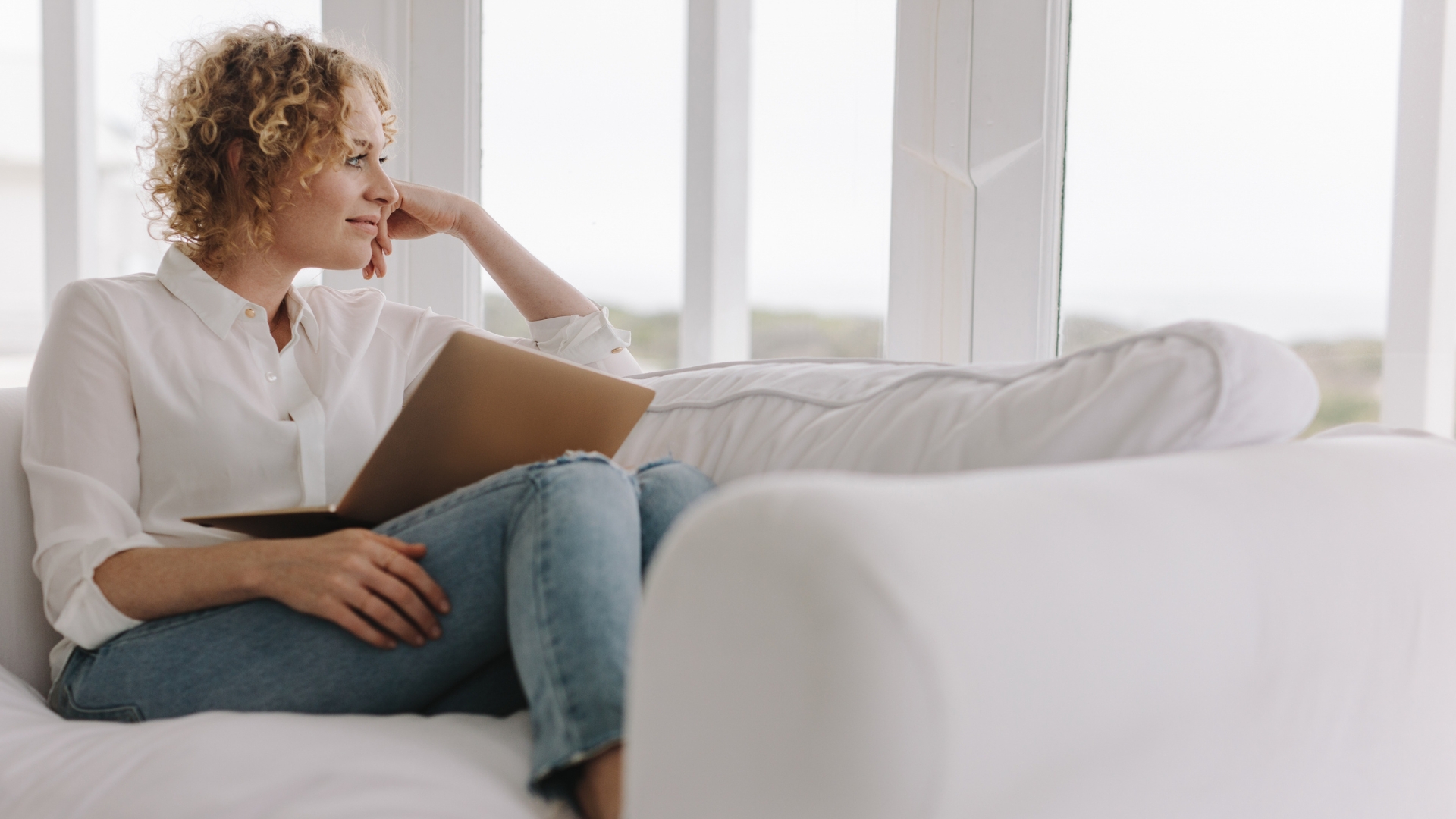 Kvinna sitter i soffan med sin laptop i knäet och ser nöjd och fundersam ut