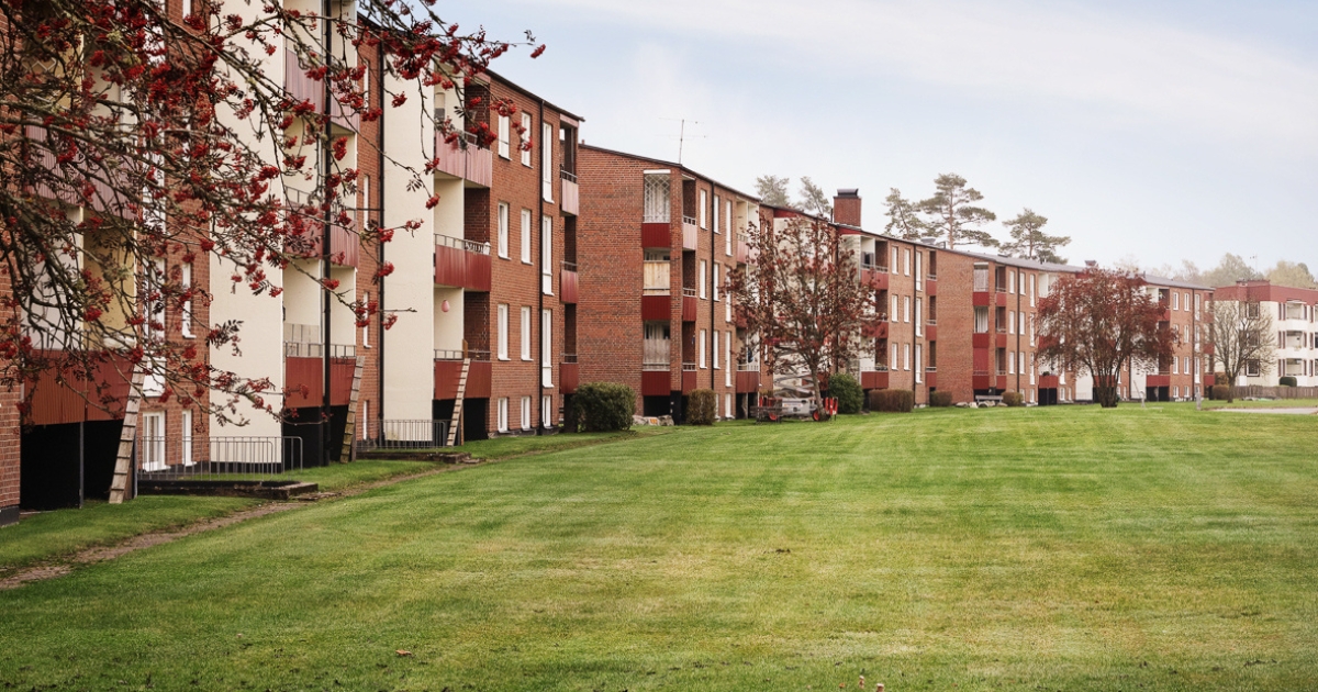 Flerbostadshus i rött tegel med balkonger i Vaggeryd, belägna längs ett stort öppet grönområde, med träd i höstfärger och barrskog i bakgrunden.