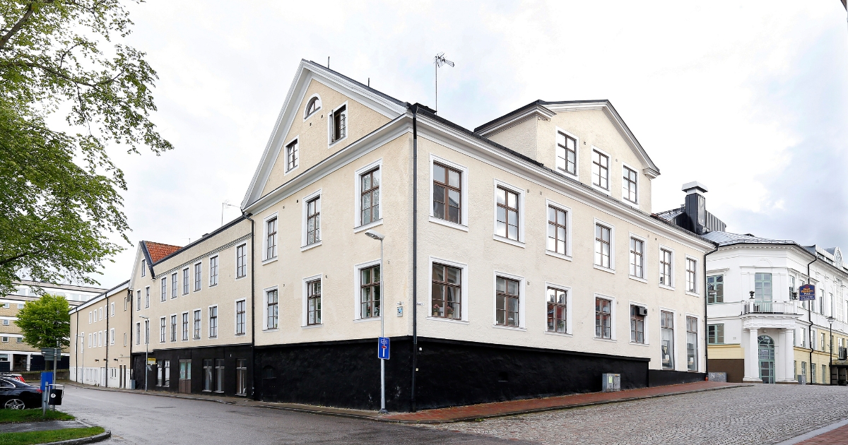 Ljusputsad hörnfastighet i centrala Västervik med klassisk arkitektur, fönster med spröjs och svart sockelvåning, belägen vid en kullerstensgata.